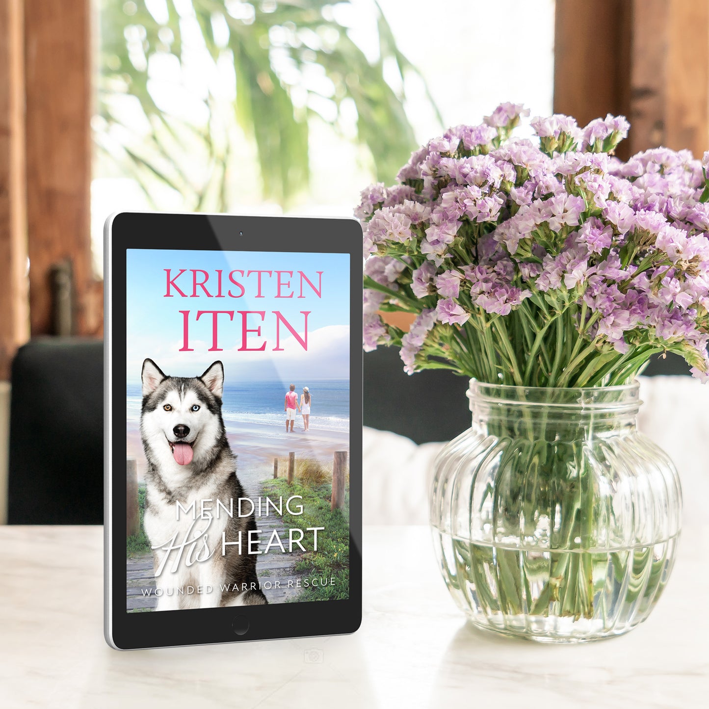 Mending his heart on an ereader sitting on a table next to a vase of flowers.